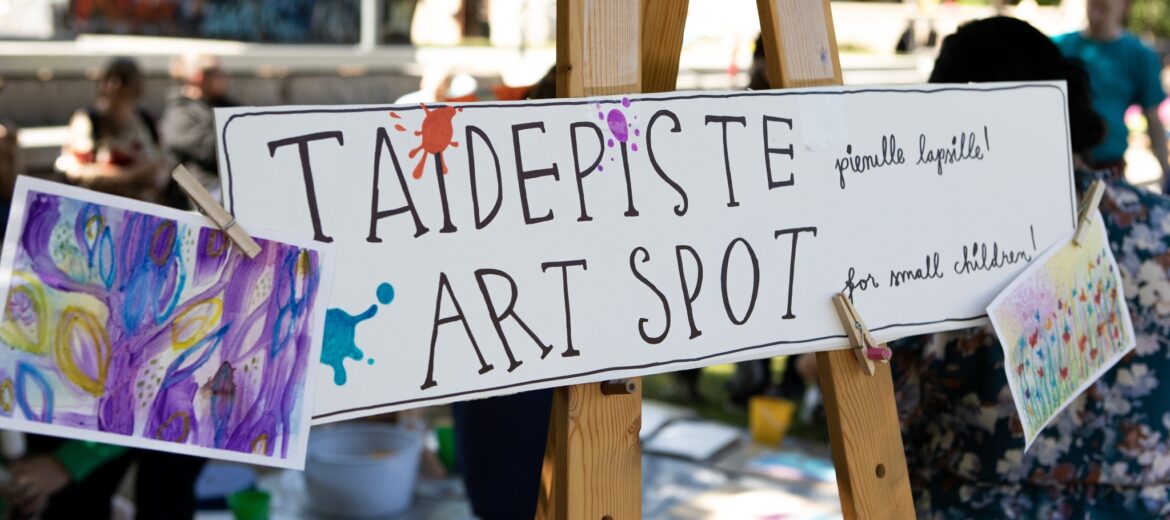 Kuvassa on maalaustelineellä kyltti, jossa teksti: Taidepiste -Art spot. Taustalla ihmisiä ulkotilassa.