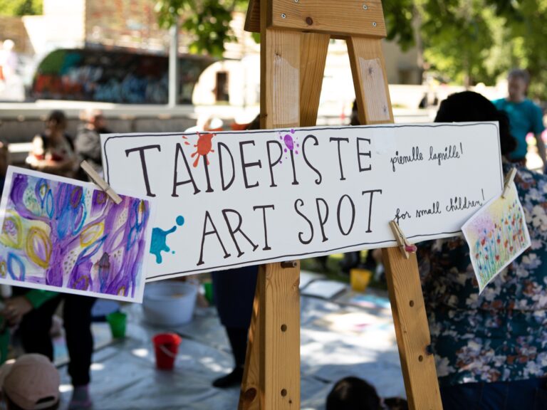Kuvassa on maalaustelineellä kyltti, jossa teksti: Taidepiste -Art spot. Taustalla ihmisiä ulkotilassa.
